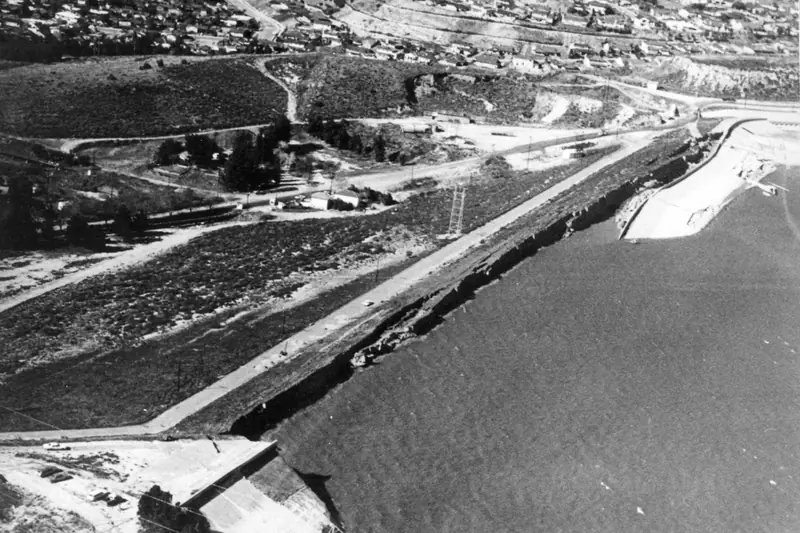 Black-and-white aerial photo of an earth dam slumped along its crest next to its reservoir, with the flat valley stretching away behind